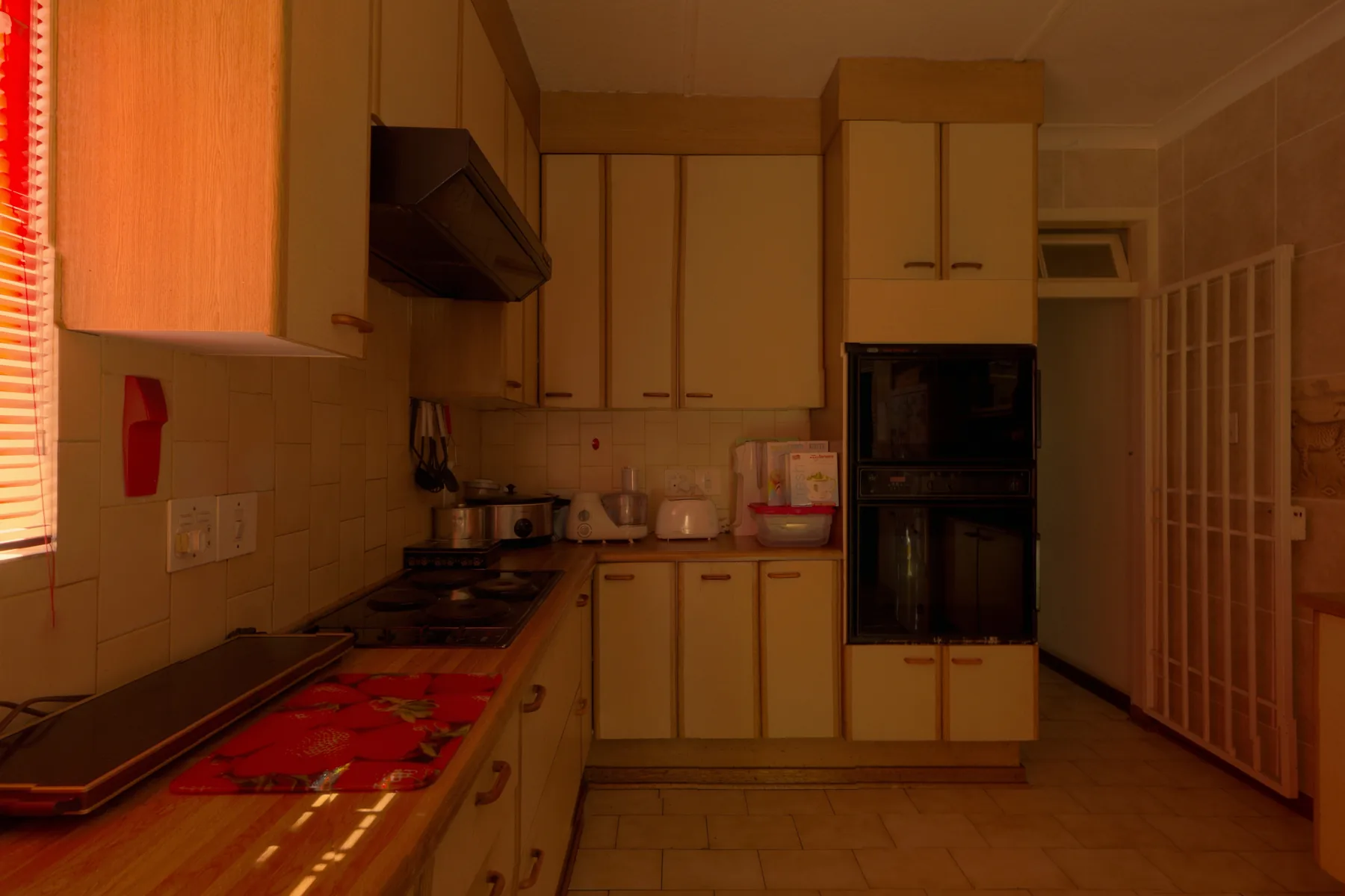 A dark kitchen lit red through the blinds, the counter full of appliances and a red mat.