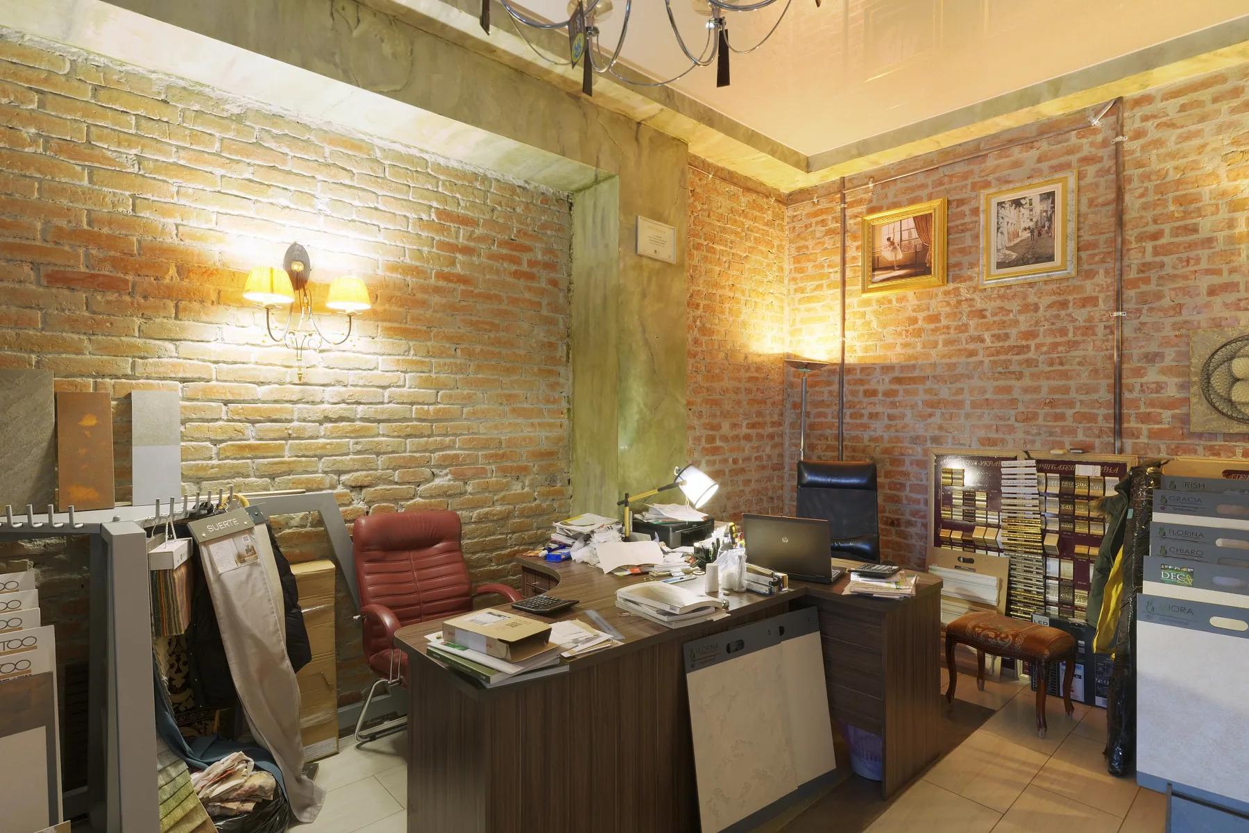 An office with a desk buried in papers, binders, a box, a mug and bottles, and hot spots from the wall lamp.