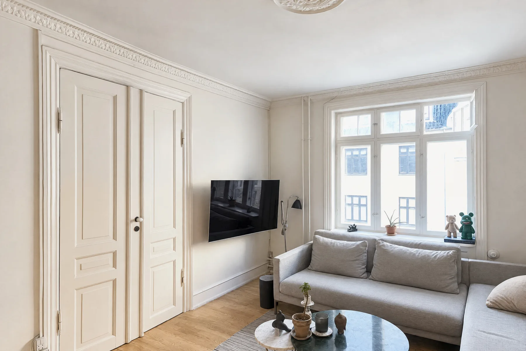Living room photographed from a corner: tall paneled double doors on the left, a wall-mounted TV, and a gray sofa under a large window, with all door frames and window edges vertical
