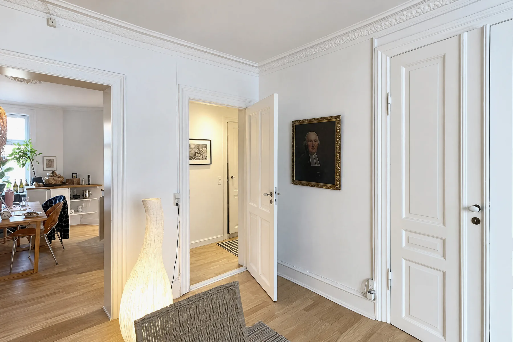 A bright living room seen toward an open doorway into a dining area and a hallway, with a tall lamp in the foreground, a framed portrait on the wall and all door frames vertical