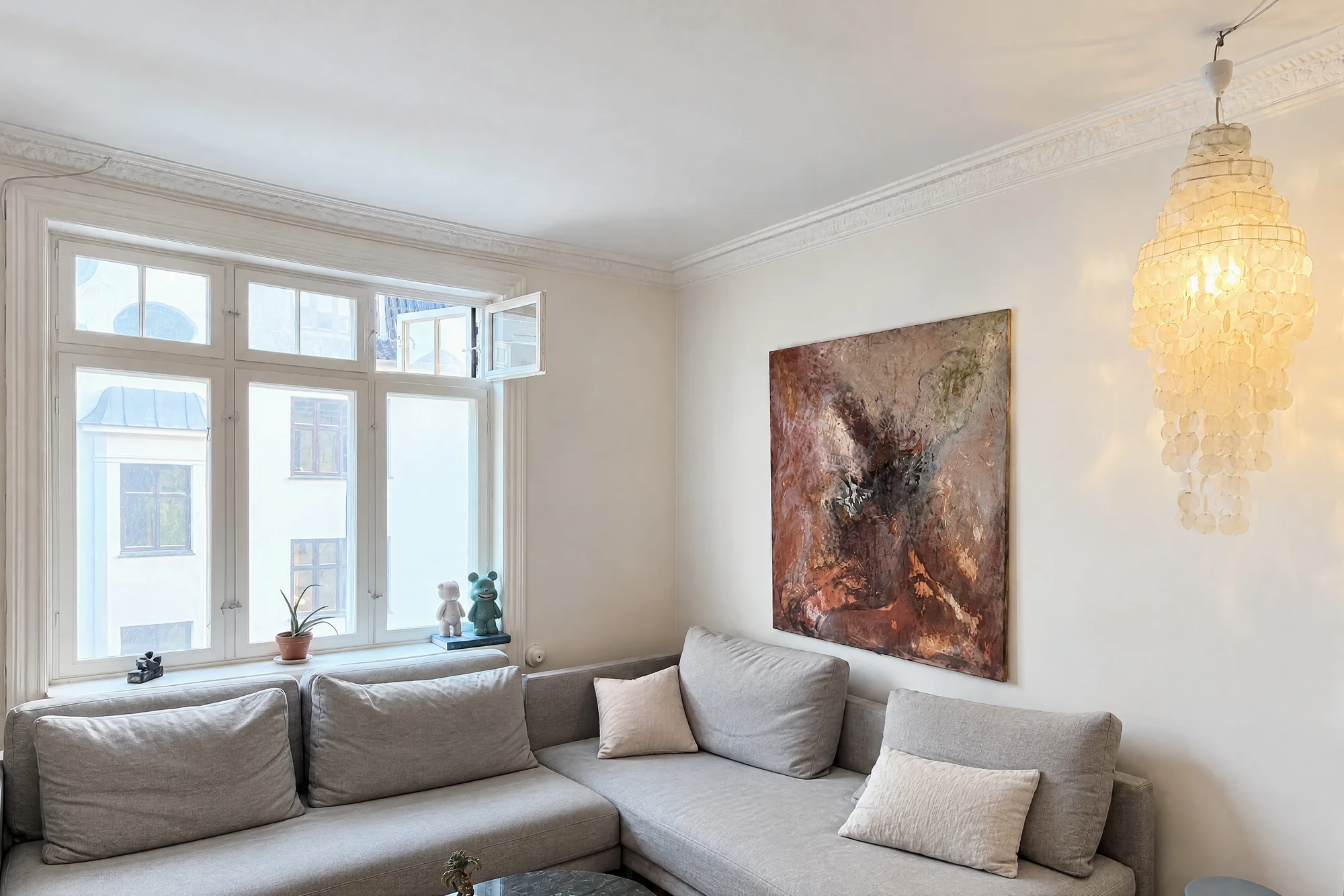 A living room with a tall paned window, a gray corner sofa with cushions, a large abstract painting in warm tones and a capiz shell pendant lamp, as October delivered it