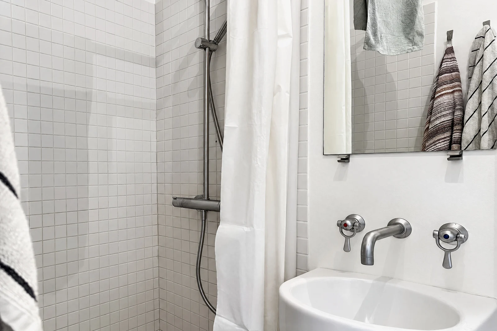 A small white-tiled bathroom: a rain shower with a white curtain, a wall-mounted basin with two taps, and a mirror reflecting striped towels on hooks