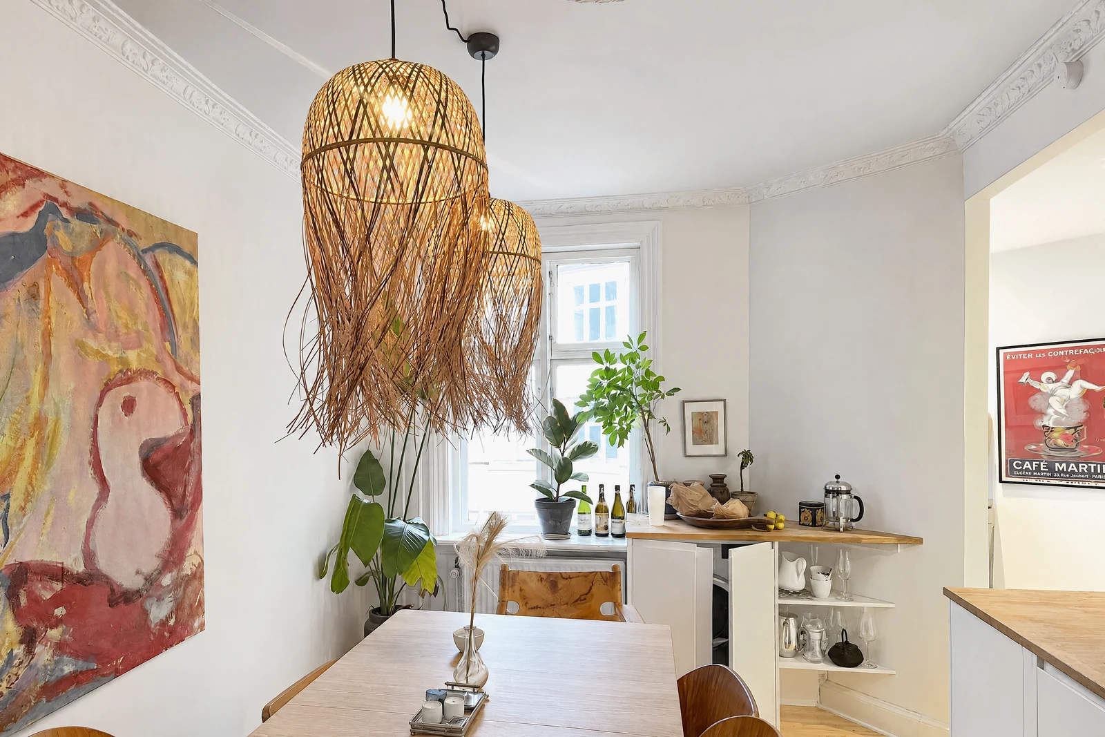 A bright dining room with two rattan pendant lamps over a wooden table, a large abstract painting, plants in a tall window and a white sideboard with glassware