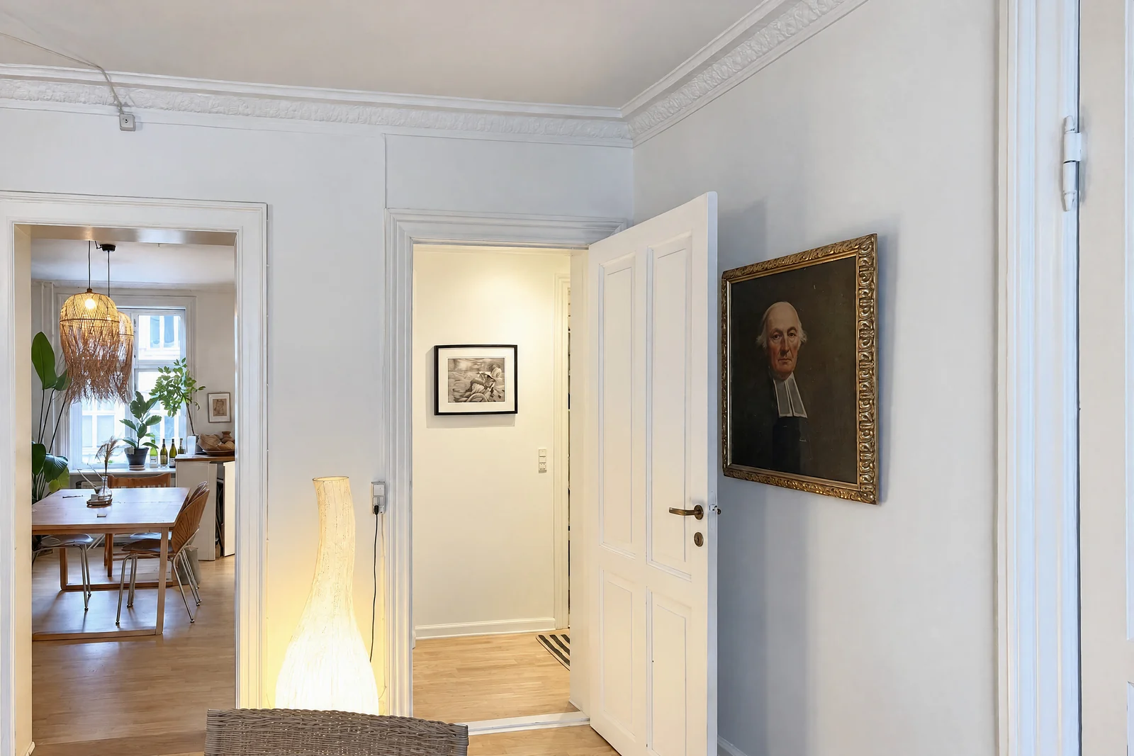 An entrance hall with white panelled doors, a gilt-framed portrait, a glowing floor lamp and a view through an open doorway into a dining room with a rattan pendant and a wooden table