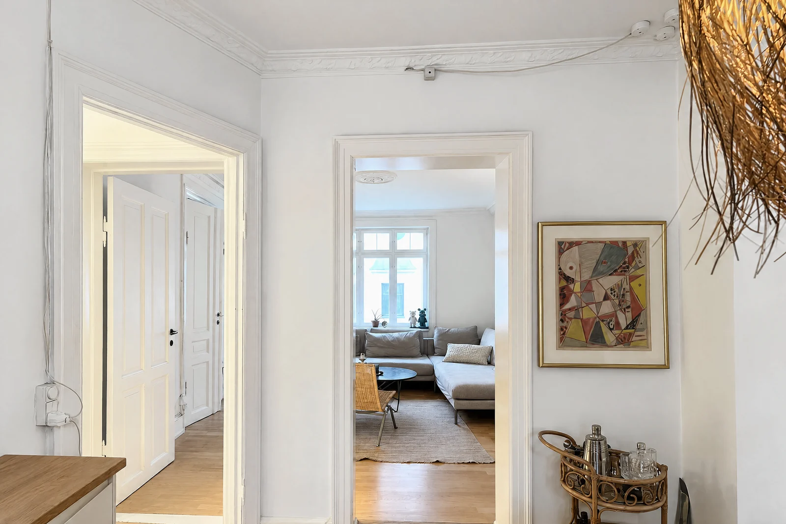 A white hallway with ornate cornicing, seen through one tall doorway into a living room with a grey sofa and a window, and through another into a corridor with white paneled doors and a wooden floor