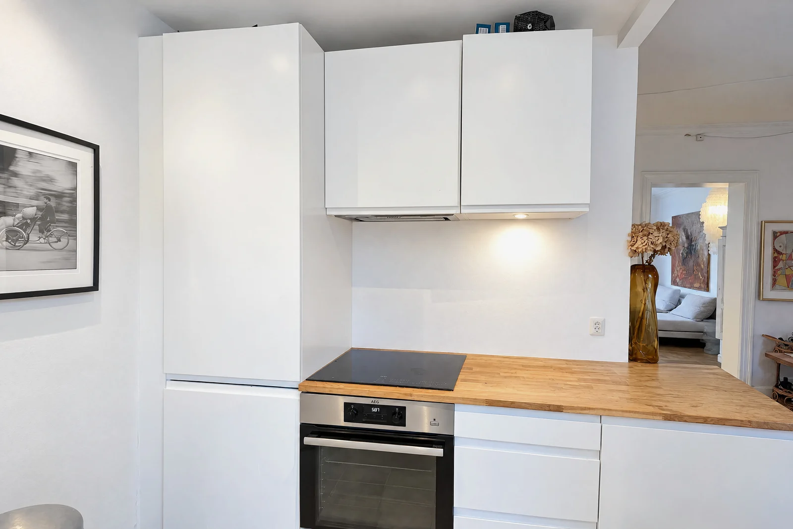 A white kitchen with flat-front cabinets, a black hob over a stainless oven, a wooden counter with only a tall amber vase of dried flowers, a framed black-and-white photo on the left wall and a doorway into a living room