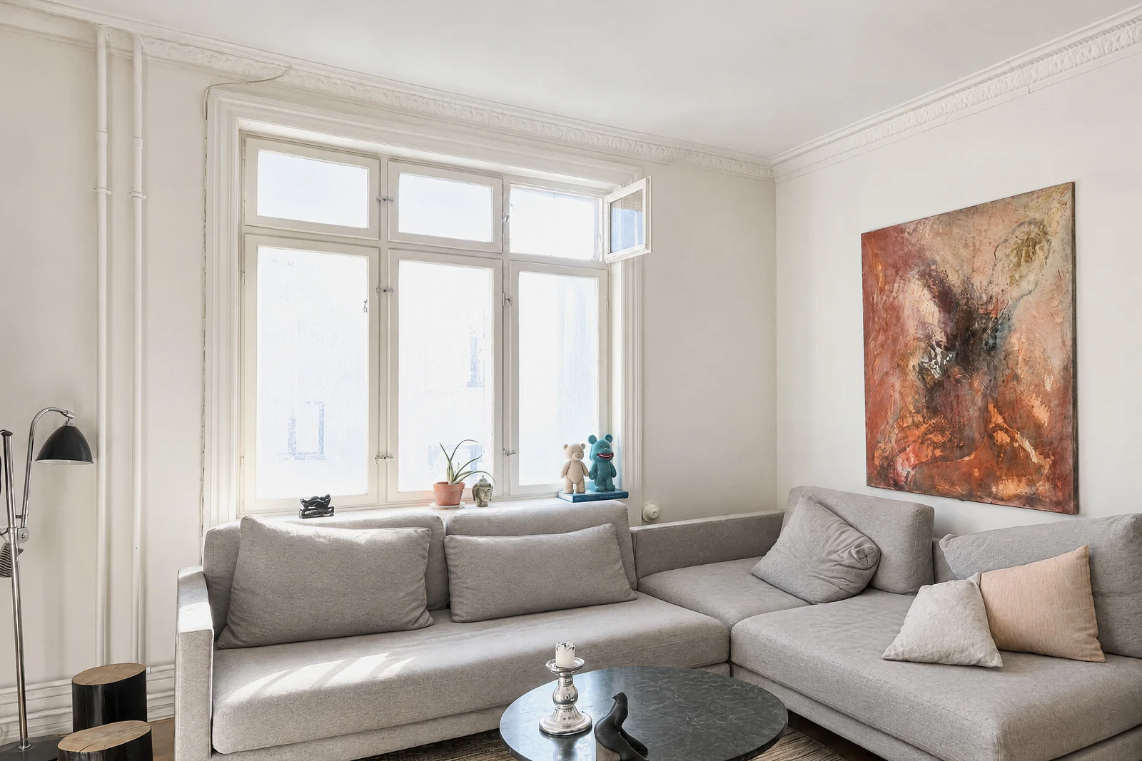 A bright living room with a grey corner sofa under a tall paned window, a round black coffee table with a candlestick, a large abstract painting in warm tones and a black floor lamp