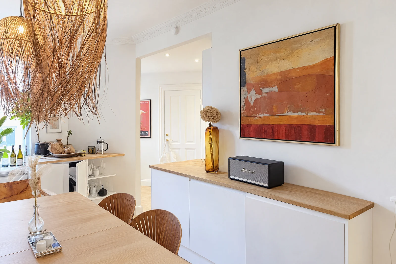 A white sideboard with an oak top under a large abstract painting in orange and red, with an amber glass vase and a black speaker, next to a wooden dining table with chairs
