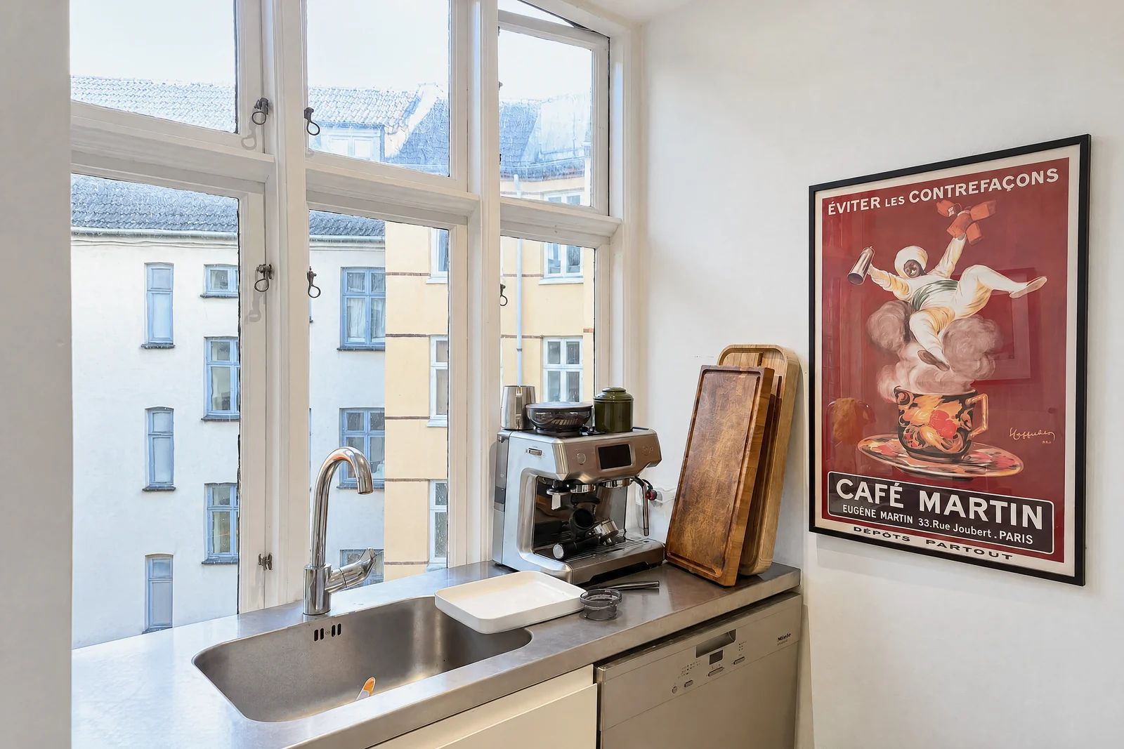 A narrow kitchen seen along the worktop: a steel sink and tap under tall paned windows with the courtyard buildings outside, an espresso machine, cutting boards and a framed Café Martin poster on the end wall